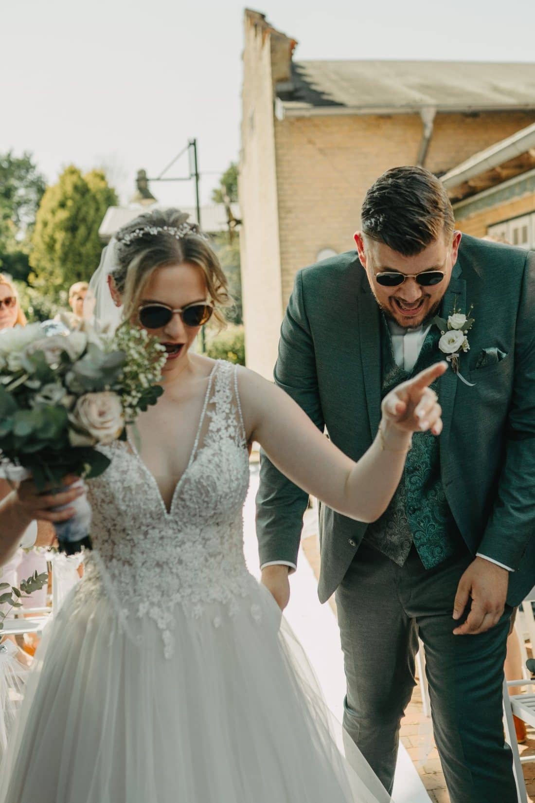 Emma und Dennis mit Sonnenbrillen nach ihrer Trauung, Emma zeigt lachend nach vorn