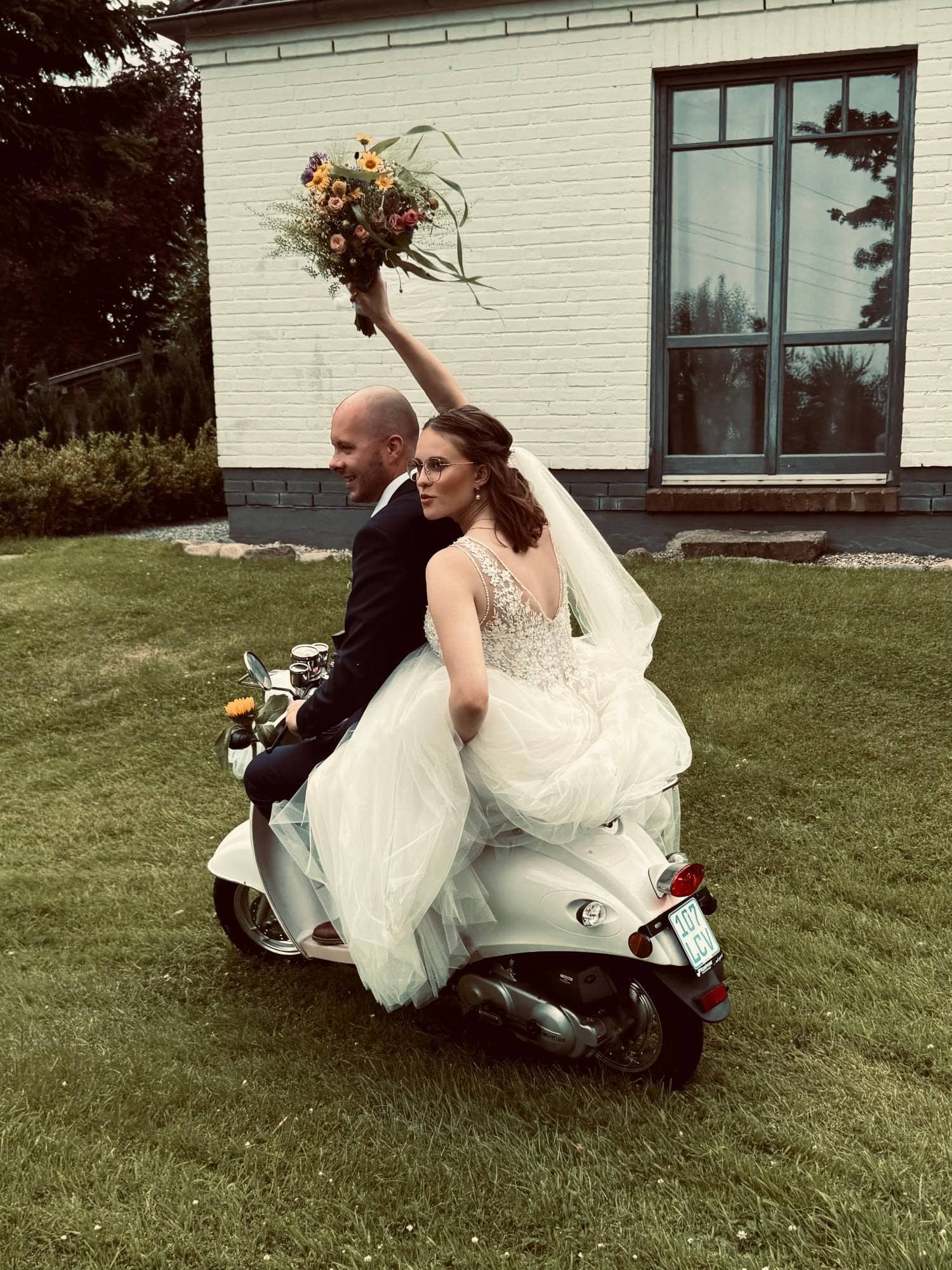 Brautpaar fährt auf einem weißen Motorroller über die Wiese, die Braut hält den Brautstrauß in die Höhe