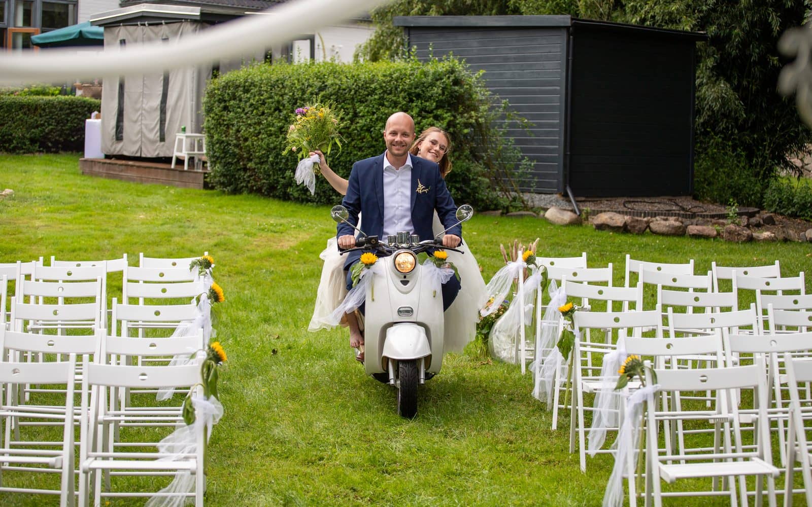 Brautpaar fährt auf einer Vespa zwischen weißen Stuhlreihen durch den Garten